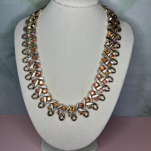 May include: A vintage gold-toned necklace with iridescent orange and pink rhinestones. The necklace is displayed on a white mannequin bust. The design features a series of interconnected loops and sparkling stones, creating a glamorous look.