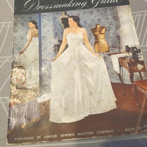 May include: A vintage Singer Dressmaking Guide booklet featuring a cover illustration of a person in a white strapless gown standing before a mirror, with a dress form and sewing machine visible in the background. The text reads Singer Dressmaking Guide.