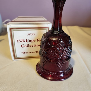 May include: A deep red glass bell with a textured diamond pattern and a smooth handle. Behind the bell sits a white cardboard box with gold trim, featuring the text AVON 1876 Cape Cod Collection Hostess Bell printed in dark red lettering.