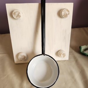 May include: A black and white enamel ladle with a long handle and a round bowl, positioned in front of a square, off-white wooden stand featuring four small, rounded feet at the corners. The ladle has a smooth, glossy finish with a black rim.