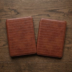 May include: Two brown leather tablet cases with a crocodile skin texture are displayed on a wooden surface. The cases are rectangular with rounded corners and a stitched border. The cases are a warm, medium brown color.