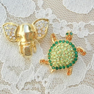 May include: Two gold-tone brooches, one shaped like an elephant and the other like a turtle. The elephant brooch is decorated with clear rhinestones, while the turtle brooch is decorated with green and orange rhinestones.