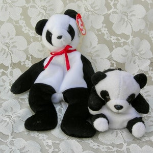 May include: Two black and white panda bear plush toys. The larger panda is wearing a red ribbon around its neck. The smaller panda is sitting in front of the larger panda.