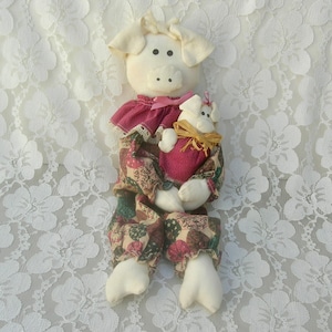 May include: A handmade pig doll dressed in a pink and brown floral print dress, holding a smaller pig doll in its arms. The dolls are sitting on a white lace background.