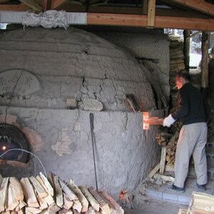 May include: A person stoking a wood-fired kiln with a long piece of wood. The kiln is made of clay and has a round opening at the bottom. There is a pile of wood in front of the kiln.