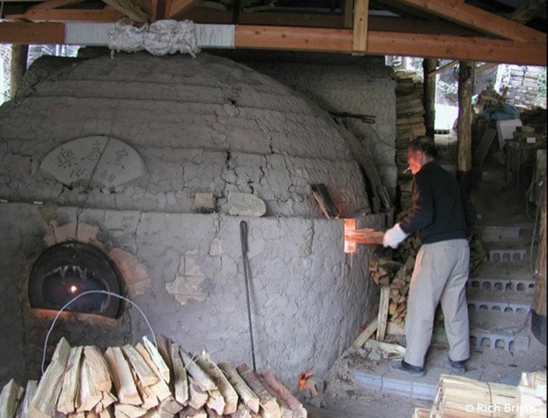 May include: A person stoking a wood-fired kiln with a long piece of wood. The kiln is made of clay and has a round opening at the bottom. There is a pile of wood in front of the kiln.