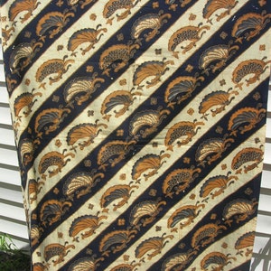 May include: A white and black fabric with a diagonal pattern of brown and gold stylised peacocks. The fabric has a traditional batik design.