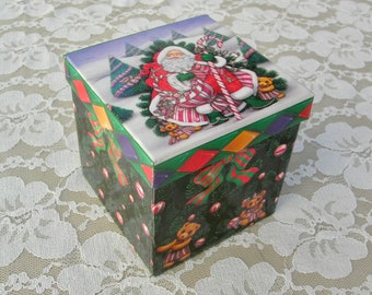 Peut inclure: Une boîte cadeau carrée sur le thème de Noël. La boîte est recouverte de papier vert avec un ruban rouge et vert. Le dessus de la boîte présente une illustration colorée du Père Noël avec une canne en sucre, entouré d'ours en peluche et de sapins de Noël.
