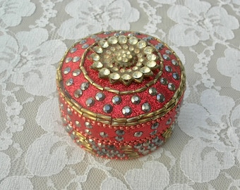 May include: A round red trinket box with a gold trim and a centre design of clear rhinestones. The box is covered in glitter.