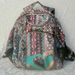 May include: A multi-colored backpack with a geometric pattern in shades of gray, pink, and teal. The backpack has multiple compartments, a top handle, and side mesh pockets. A logo patch is on the front. The backpack is designed for carrying school supplies or personal items.