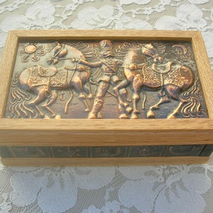 May include: Rectangular wooden box with a copper-colored relief depicting a person with two horses. The relief is framed by a light-colored wood. The box has a hinged lid and is likely used for storage or display.