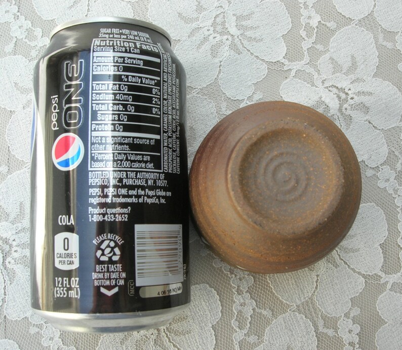 May include: A black can of Pepsi One cola with the nutrition facts listed on the side. The can is next to a small, brown ceramic bowl.