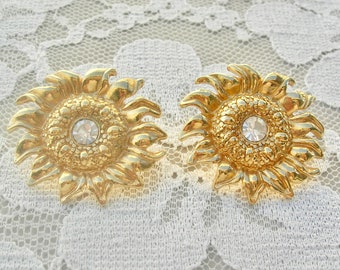 May include: A pair of gold-coloured sunflower earrings. Each earring has a central clear rhinestone and detailed petal design. The earrings are set against a white lace background.