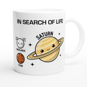 May include: White ceramic mug with the text "IN SEARCH OF LIFE" and illustrations of Saturn, Enceladus, and Titan. Saturn is depicted with a smiling face and rings. The mug is a standard size.