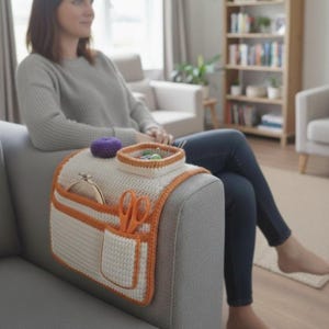 May include: A cream-colored crochet sofa arm organizer with orange trim and pockets. It holds a purple pincushion, a small basket with sewing supplies, an embroidery hoop, and orange scissors. The organizer is designed to keep craft supplies organized and accessible.