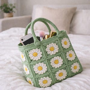 Op de afbeelding: Een groene gehaakte tote bag met witte madeliefjes applicaties. De tas heeft twee handvatten en is gevuld met een smartphone, make-up borstels en andere schoonheidsproducten. De tas ligt op een wit sprei.