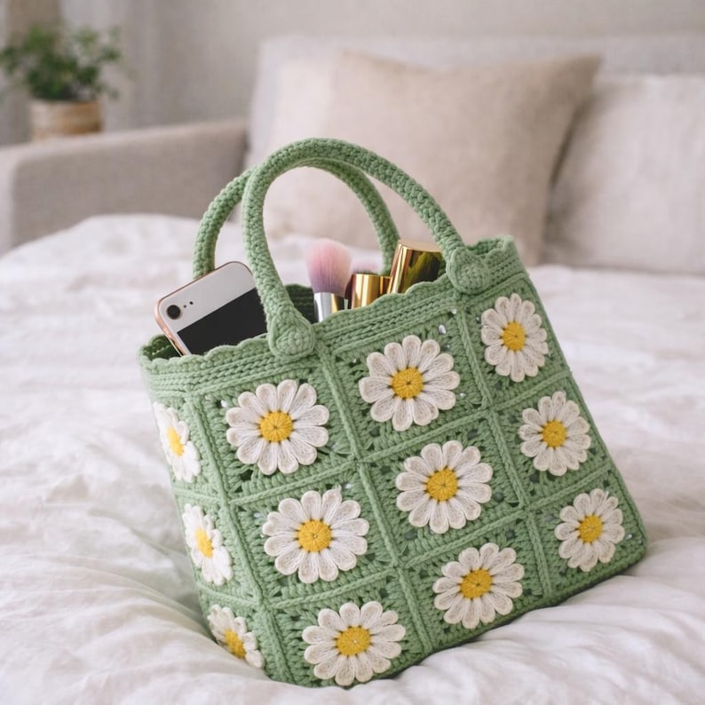 Op de afbeelding: Een groene gehaakte tote bag met witte madeliefjes applicaties. De tas heeft twee handvatten en is gevuld met een smartphone, make-up borstels en andere schoonheidsproducten. De tas ligt op een wit sprei.
