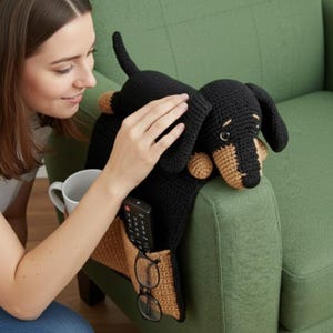 May include: A black and tan crochet dachshund-shaped armrest organizer with pockets for a remote, glasses, and a mug. The dog is lying on a green sofa arm. The organizer is handmade.
