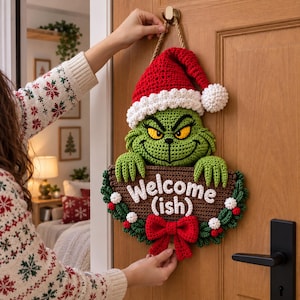 Puede incluir: Un colgante de pared de ganchillo con un personaje verde que lleva un gorro de Papá Noel rojo y blanco, sosteniendo un cartel marrón con el texto blanco Welcome (ish). El cartel está decorado con hojas de acebo, bayas blancas y un lazo rojo.
