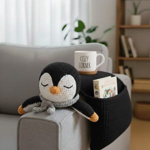Può includere: Un pinguino all'uncinetto con una sciarpa grigia e gli occhi chiusi poggia su un bracciolo grigio. Un organizer per braccioli nero contiene un libro e una tazza bianca con la scritta "Cozy Corner". Il pinguino ha dettagli neri, bianchi e arancioni.