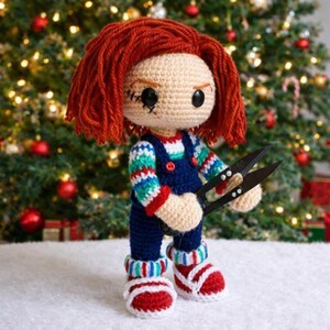 May include: Crocheted doll resembling a character with red hair, holding black scissors. The doll wears a blue jumpsuit, striped sleeves, and red and white striped shoes. The background features a Christmas tree with ornaments.