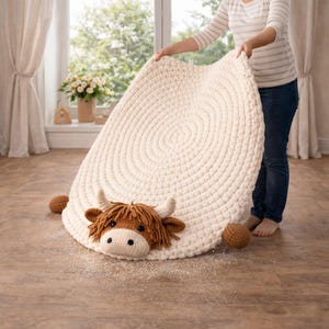 May include: A cream-colored, round, crocheted rug with a Highland cow design. The cow has brown fur, horns, and a white face. Two brown balls are on either side of the cow. The rug is approximately 48 inches in diameter.