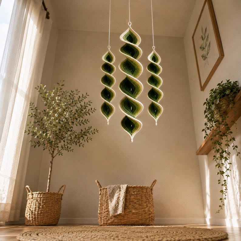 May include: Three hanging spiral ornaments made of crocheted yarn in shades of cream and green. The central ornament is larger than the two flanking it, and all three are suspended by thin white strings from an unseen ceiling point.