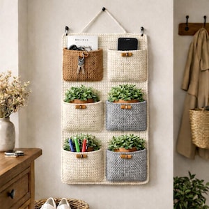 May include: A wall-mounted organizer with six pockets in varying shades of beige and gray. The pockets hold keys, a phone, pens, and small potted plants. The organizer is made of woven material and hangs from a rope.