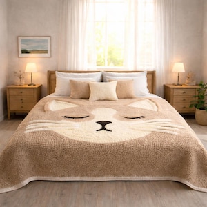 May include: A beige and cream-colored bedspread with a large cat face design. The cat's eyes are closed, and the nose is black. The bed is made with pillows and a wooden bed frame. The room has two nightstands and lamps.