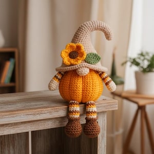 Può includere: Una bambola a uncinetto fatta a mano a forma di gnomo con un corpo a forma di zucca. Lo gnomo indossa un cappello beige adornato con un girasole giallo e una foglia verde. Le sue braccia e gambe hanno motivi a strisce e le gambe penzolano. La bambola è seduta su una superficie di legno.