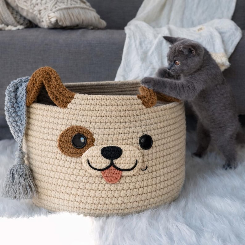 May include: A beige crochet dog-faced basket with brown ears and a brown spot. The basket has a black nose, eyes, and a pink tongue. A gray kitten is playing with the basket.