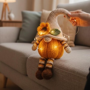 May include: A crocheted pumpkin gnome with a beige hat, orange body, and striped legs. The gnome has a flower and leaf on its hat and is illuminated by small lights. The gnome is sitting on a gray couch.