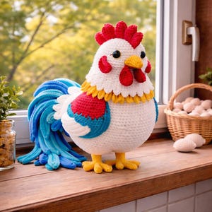 May include: A handmade crochet rooster with a white body, red comb, and yellow beak and feet. The rooster has a blue and white tail and is standing on a wooden surface. A basket of eggs is in the background.