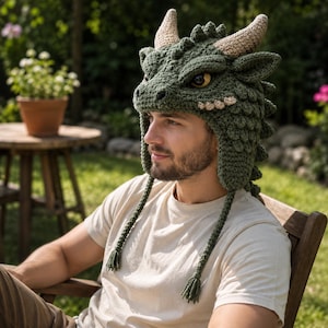 May include: A dark green crocheted hat shaped like a dragon head, featuring two light tan horns, textured scales, and large yellow eyes. The hat includes ear flaps with long braided tassels hanging down on each side of the head.