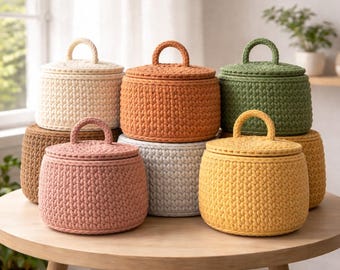May include: A selection of crochet storage baskets in shades of cream, orange, green, pink, grey, and yellow. Each basket has a matching lid and a small handle. The baskets are displayed on a round wooden table.