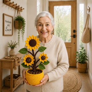 Puede incluir: Una planta de girasol de ganchillo con tres flores de color amarillo brillante con centros marrón oscuro, hojas verdes y un tallo marrón, en una maceta de ganchillo amarilla texturizada. La planta es sostenida por una persona con cárdigan beige.