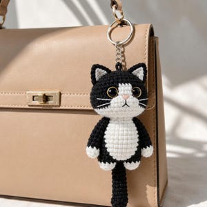 May include: A handmade black and white crochet cat keychain with a silver keyring. The cat has large, round eyes, a pink nose, and white whiskers. The keychain is hanging from a beige handbag.