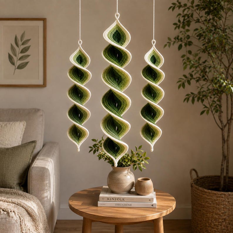 May include: Three hanging spiral decorations crafted from yarn in an ombre gradient of cream, light green, and dark forest green. Each piece features a twisted, elongated shape suspended by a thin white string, showcasing a textured crochet stitch.