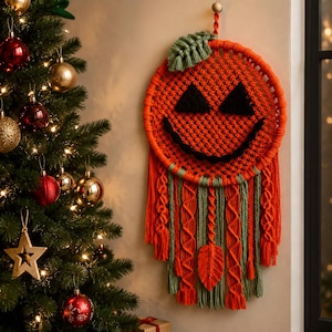 May include: A circular macrame wall hanging shaped like a jack-o'-lantern, featuring orange cord with black triangular eyes and a curved mouth. The piece includes a green leaf at the top, hanging orange and green fringe, and a central leaf accent.