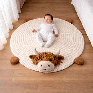May include: A cream-colored, round, crocheted rug with a Highland cow head and four brown crocheted balls. A baby in a white onesie is lying on the rug. The cow head has brown fur, horns, and a smiling face.