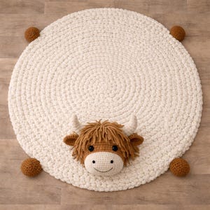 May include: A round, off-white crochet rug with a Highland cow face in the center. The cow has brown fur, horns, and a smiling face. Four small brown pom-poms are attached to the rug's edges.