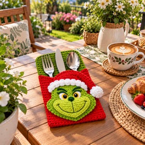 Puede incluir: Un porta cubiertos de ganchillo en verde y rojo con la cara de un personaje verde sonriente que lleva un gorro de Papá Noel rojo con borde blanco. El soporte contiene un tenedor, un cuchillo y una cuchara de plata para organizar.
