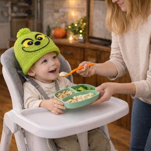 Peut inclure: Un jeune enfant portant un bonnet vert tricot&eacute; avec un visage de dessin anim&eacute; est assis dans une chaise haute. L'enfant est nourri avec une cuill&egrave;re orange et a une assiette verte divis&eacute;e avec de la nourriture.