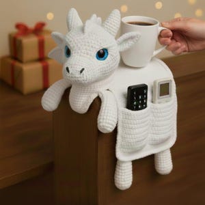 May include: A white crocheted dragon-shaped organizer with blue eyes, holding a white mug of coffee. The organizer has pockets for a remote control and other small items. The dragon is draped over a wooden surface.