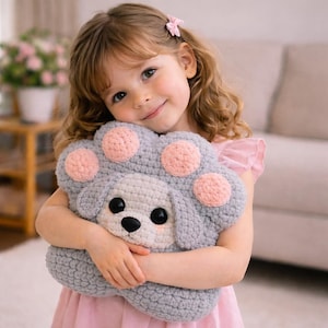 May include: A gray crocheted paw-shaped pillow with pink circles and a puppy face. The puppy has black eyes and a black nose. The pillow is soft and cuddly, perfect for a child's room or as a decorative item.