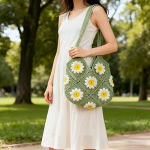 May include: A green crochet tote bag with white daisy appliqués and yellow centers. The bag has a diamond pattern and a long strap. The background is a park with green trees and grass.
