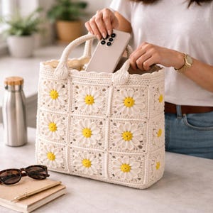 Op de afbeelding: Cr&egrave;mekleurige gehaakte tote bag met een madeliefjespatroon. De tas heeft witte bloemblaadjes en gele centra, met stevige handvatten. Een persoon plaatst een telefoon in de tas. Zonnebril, notitieboek en waterfles zijn ook zichtbaar.