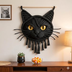 Peut inclure: Décoration murale en macramé noir représentant un chat avec des yeux jaune doré et des moustaches. Le visage du chat est fait de grosse corde noire tissée, suspendu à une cheville en bois. Un portrait de chat en noir et blanc encadré est visible.