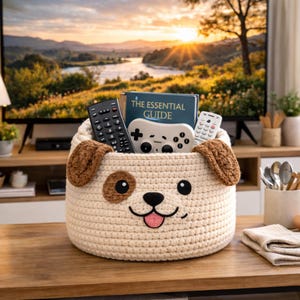May include: A cream-colored, crocheted dog-shaped basket holding a book, two remote controls, and a video game controller. The basket has brown ears, eyes, and a pink tongue. The basket is on a wooden table in front of a television.