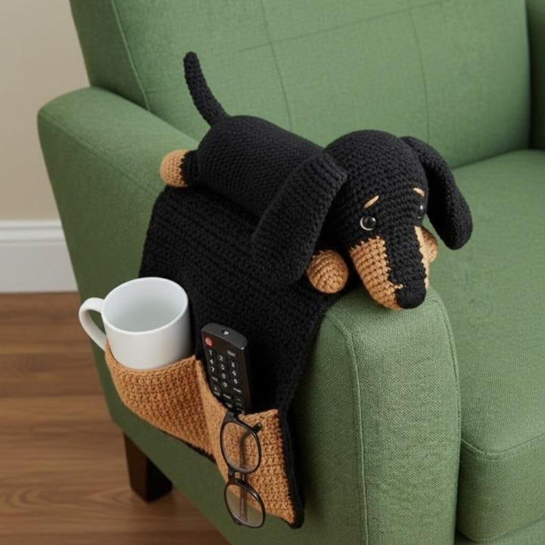 May include: A black and tan crochet dachshund armrest organizer with pockets for a remote, glasses, and a mug. The dachshund is lying on a green armchair. The organizer is a unique and functional home decor item.
